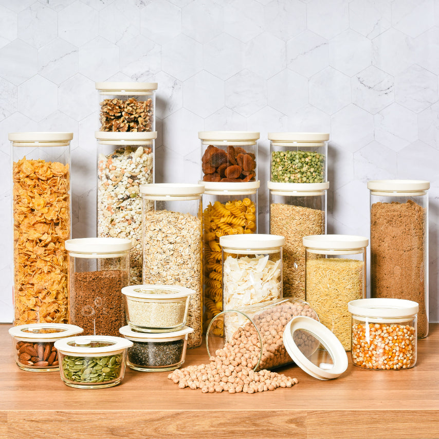 Glass pantry storage containers with airtight lids displaying colourful pasta and grains on a wooden bench.