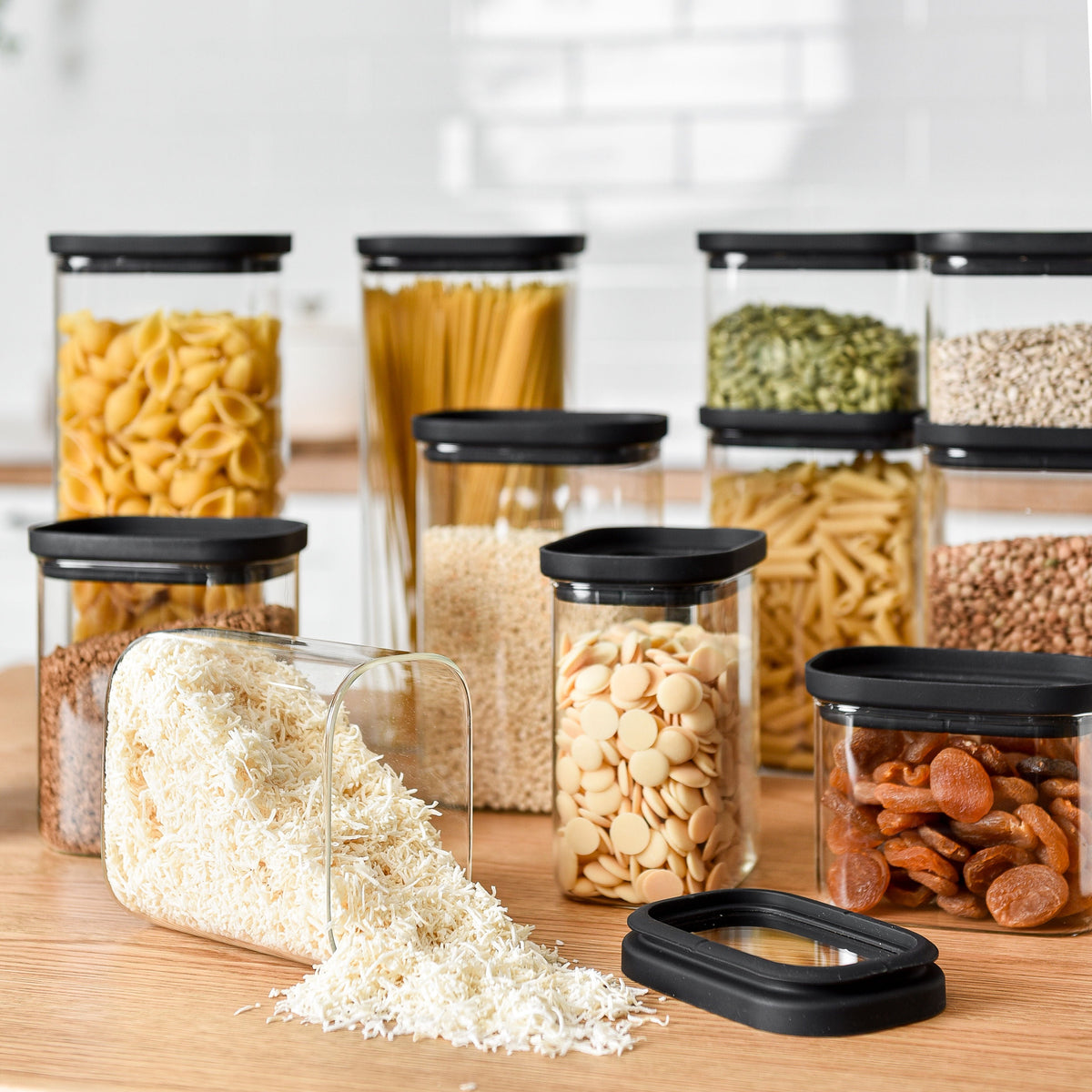 A glass pantry jar bundle with black lids storing dry goods, with one jar open on a wooden kitchen bench.
