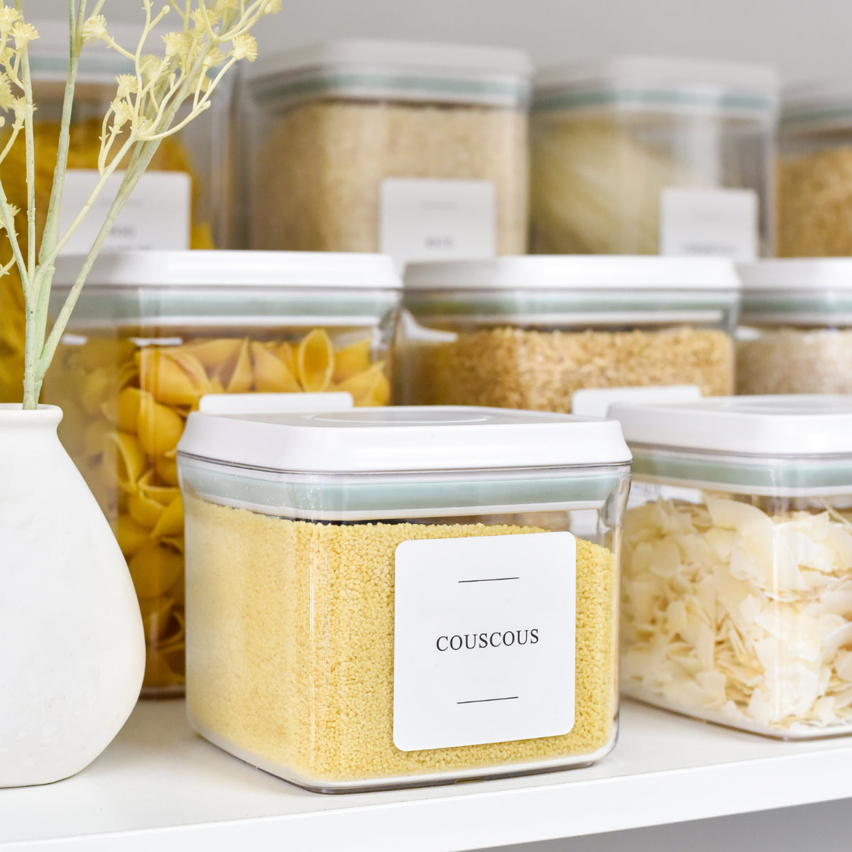 Clear pantry storage containers with white lids and labels neatly organising pasta and grains on a shelf.